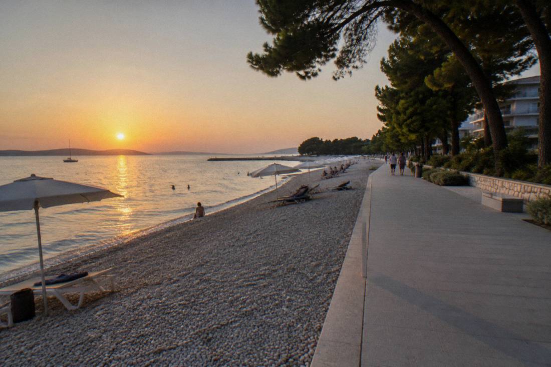 A long pebble beach at Borik lined with pine trees and a wide promenade, calm sea and a few distant swimmers
