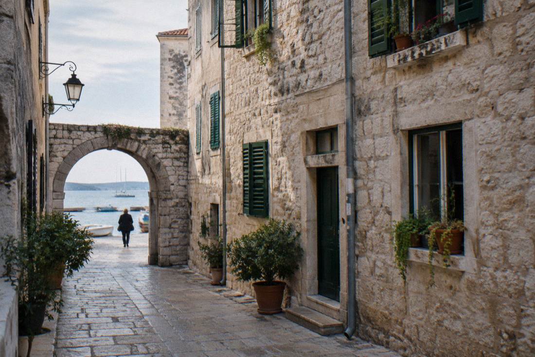A narrow limestone lane in Zadar old town under a round arch, potted plants on the windowsills and green shutters
