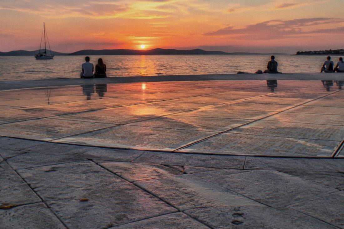 A wide circle of glass panels set in the Zadar waterfront paving at sunset, orange sky over the sea and a dark island
