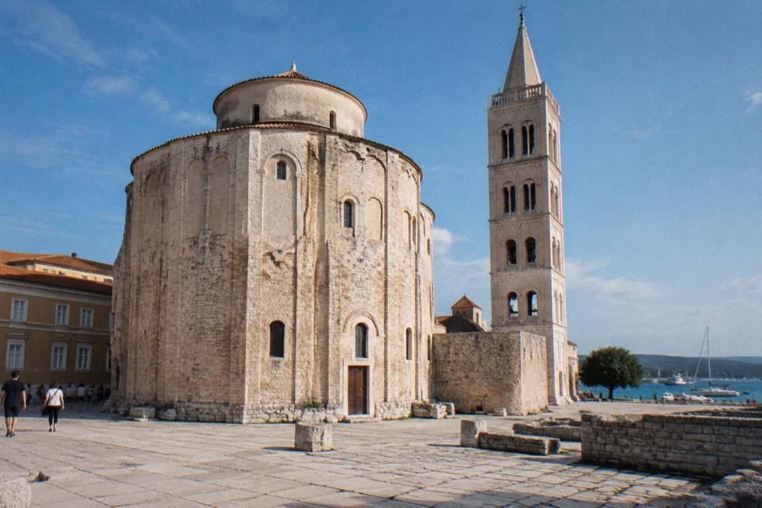 A massive round pre-Romanesque stone church with a conical roof seen across a paved square in clear morning light