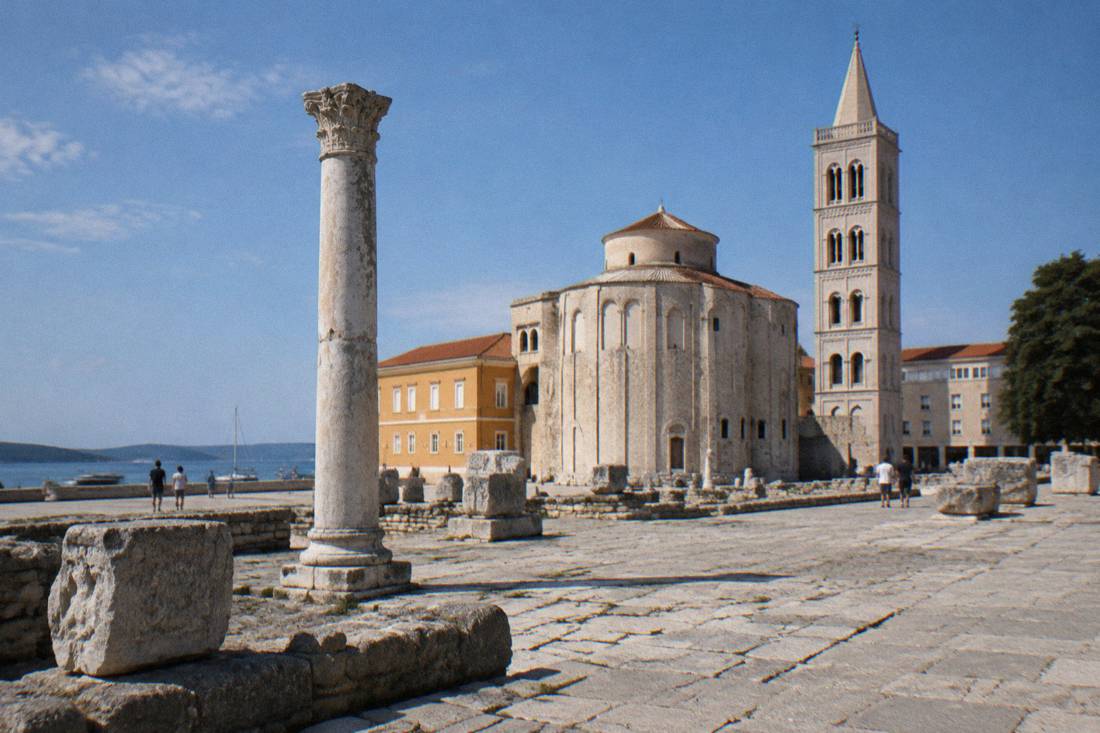 Weathered Roman forum paving with a single standing column and carved stone blocks, a round old stone church behind in morning sun