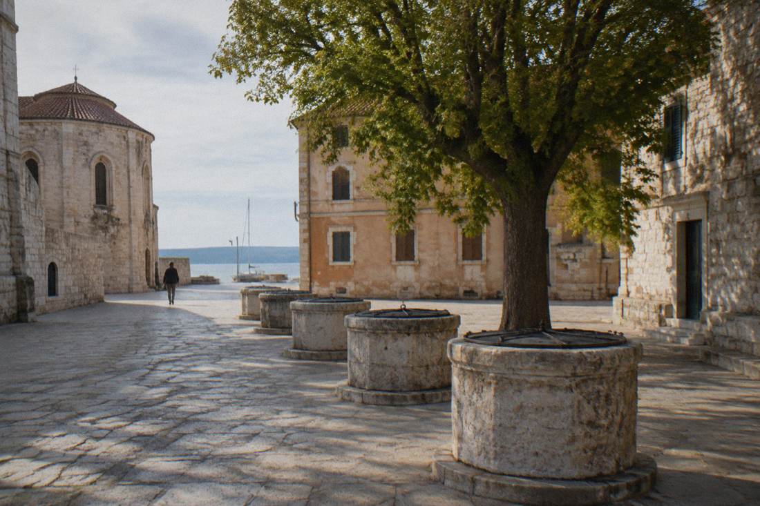 Five round stone well-heads in a row on a small shaded square surrounded by old stone buildings