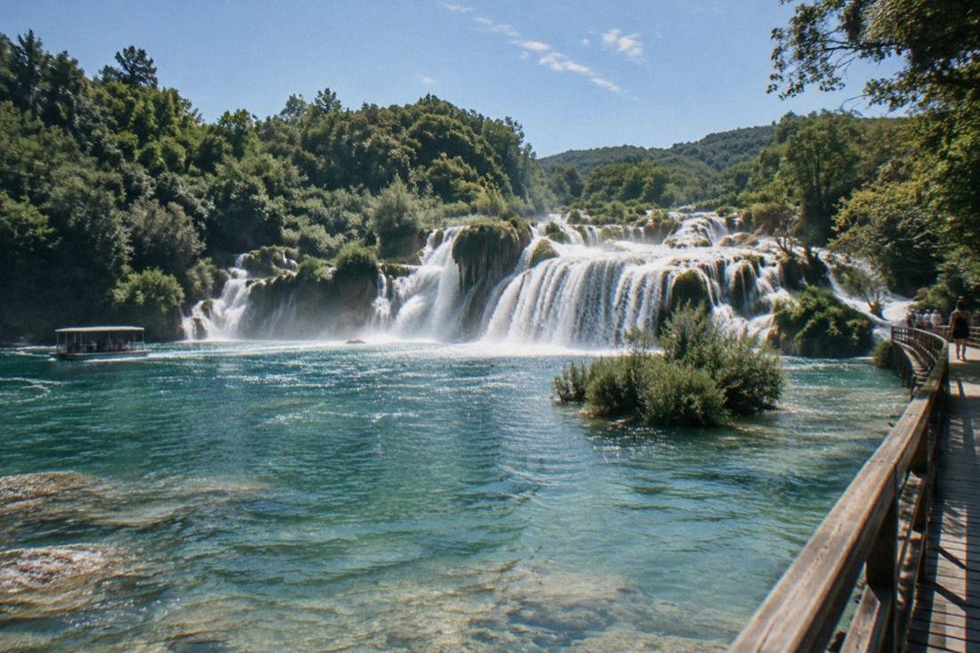 Wide tiered travertine waterfalls dropping through green forest into a turquoise pool at Krka, wooden boardwalks at the edge