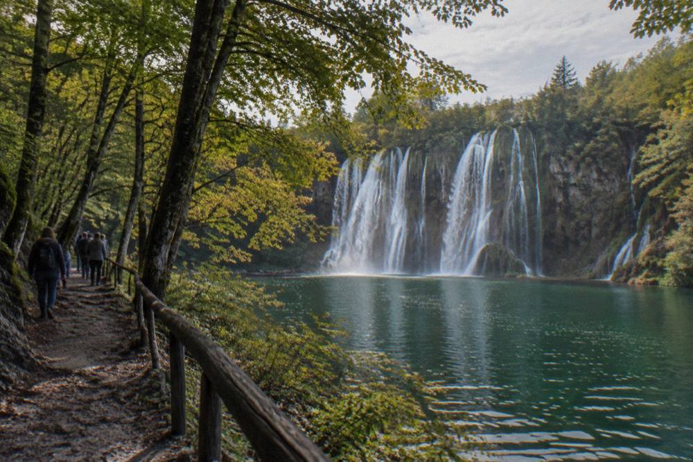 A broad waterfall dropping into a green lake beside a shaded forest path at Plitvice