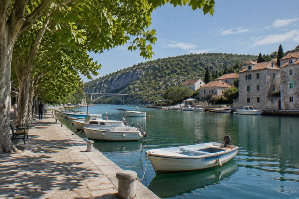 Skradin's stone quay and small moored boats on the calm river beneath shade trees
