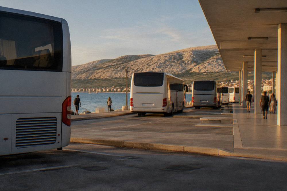 Intercity coaches parked in bays at a coastal bus station with a limestone hill behind