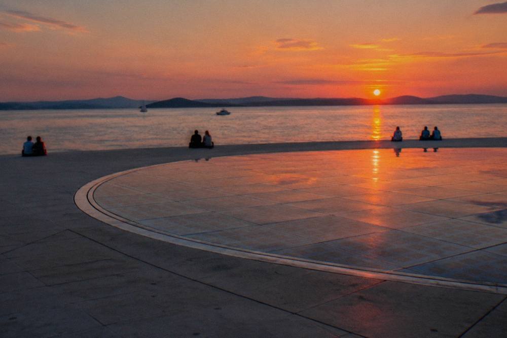 The circular stone plaza of the Greeting to the Sun at sunset with a few people far off and the sea behind