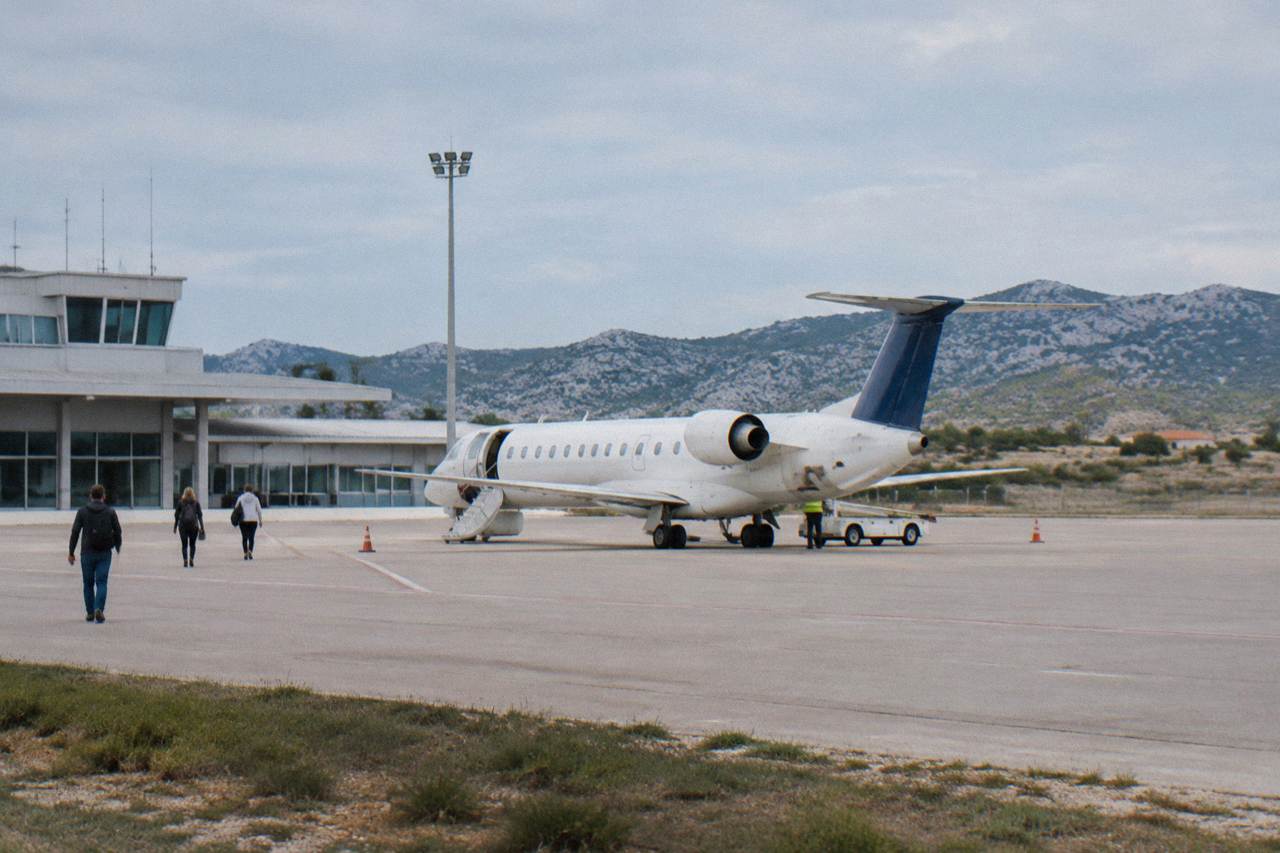 A small passenger plane on a sunlit apron near a low terminal with dry hills behind at Zadar airport