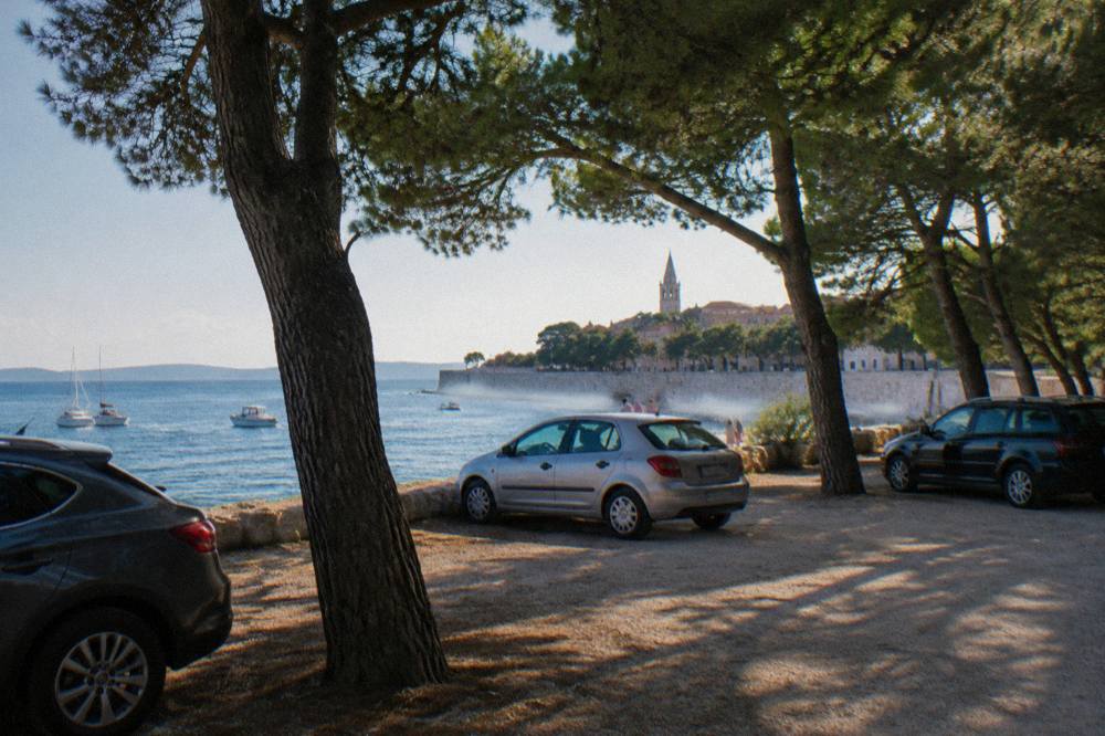 A pine-shaded car park beside Zadar's old town wall with the blue sea just beyond