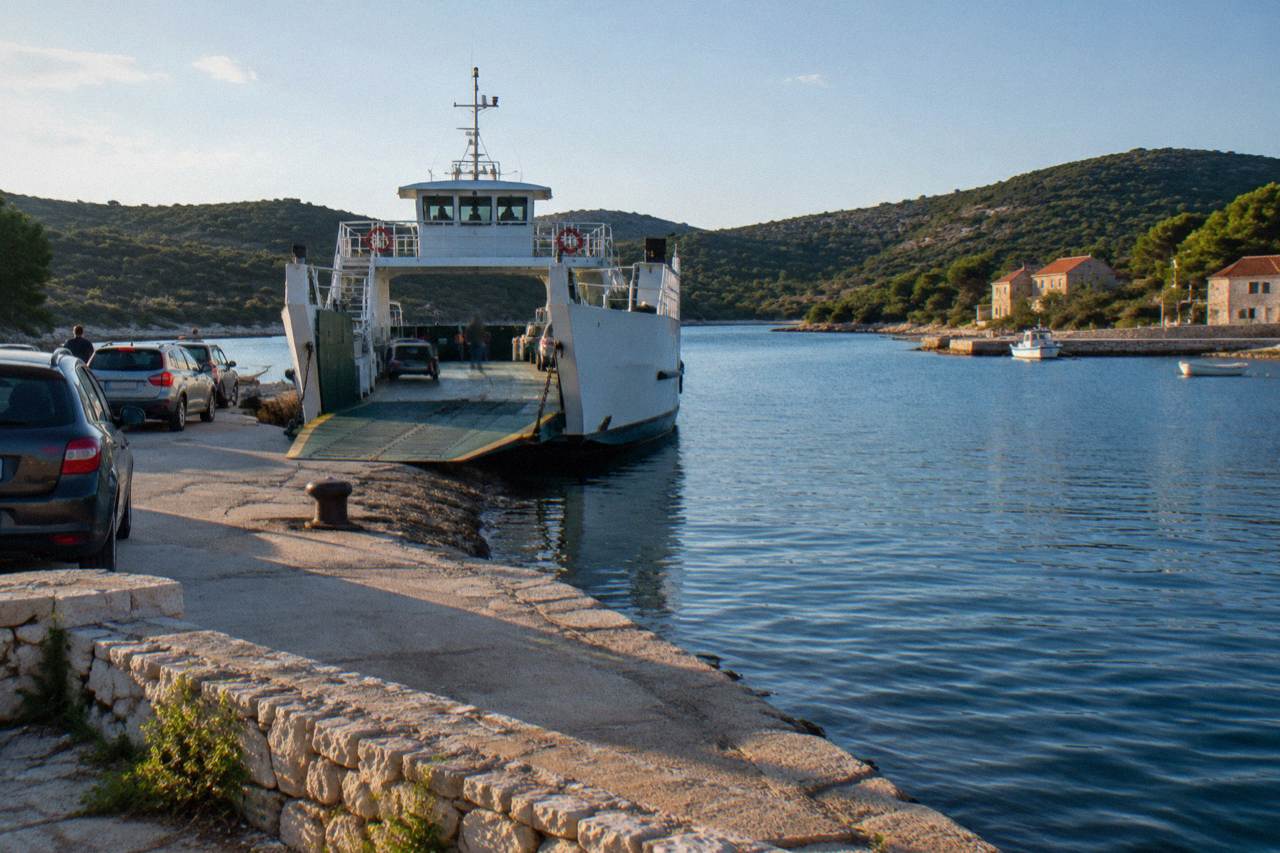 A small car ferry at the quiet stone harbour of Brbinj on Dugi Otok with scrubby pine hills behind