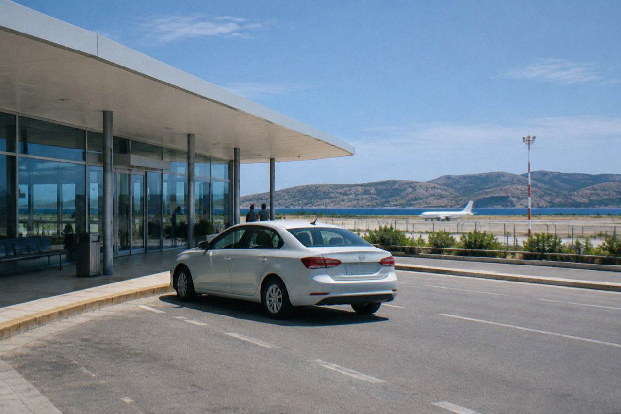 A light-coloured car waiting at the kerb outside a low airport terminal with dry hills beyond