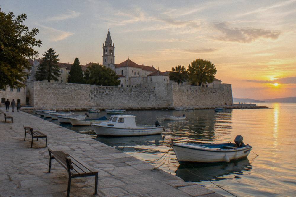 A stone quay with empty benches and small moored boats at golden hour along Zadar's old town wall