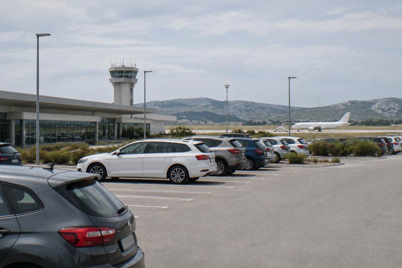 Rows of parked cars in an open car park beside a low terminal with karst hills on the horizon