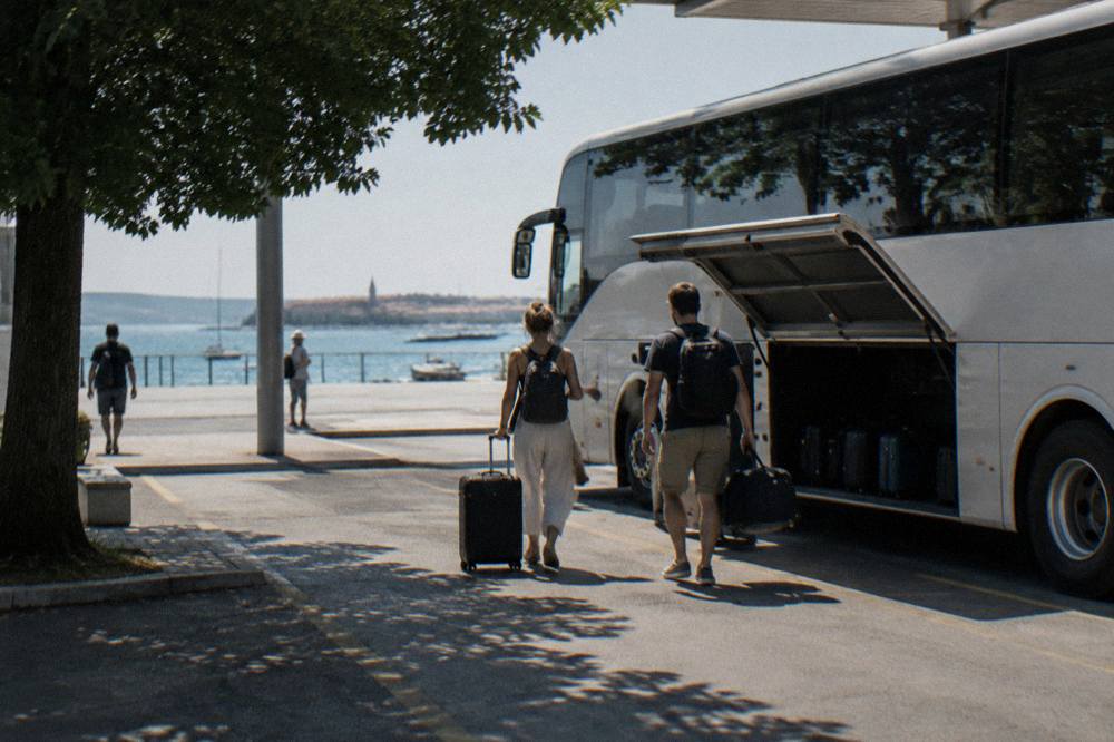 A coach with open luggage hold at a bus station bay with two travellers seen from behind