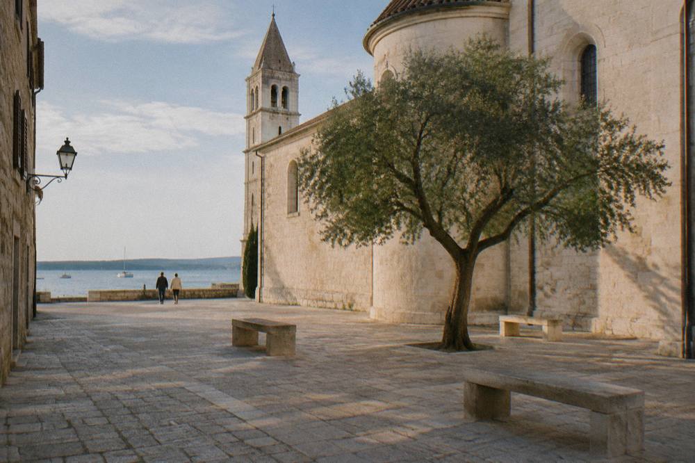 A quiet stone square with a pale church wall, a tree and empty stone benches in Zadar Old Town