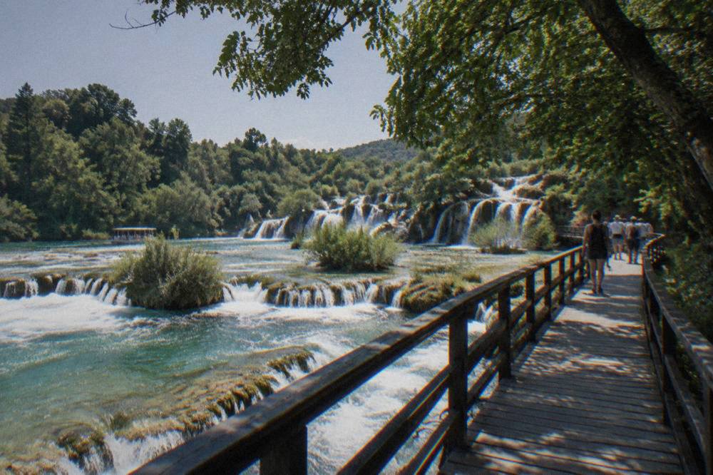 A wooden walkway crossing travertine barriers and rushing water at Krka with a few distant visitors