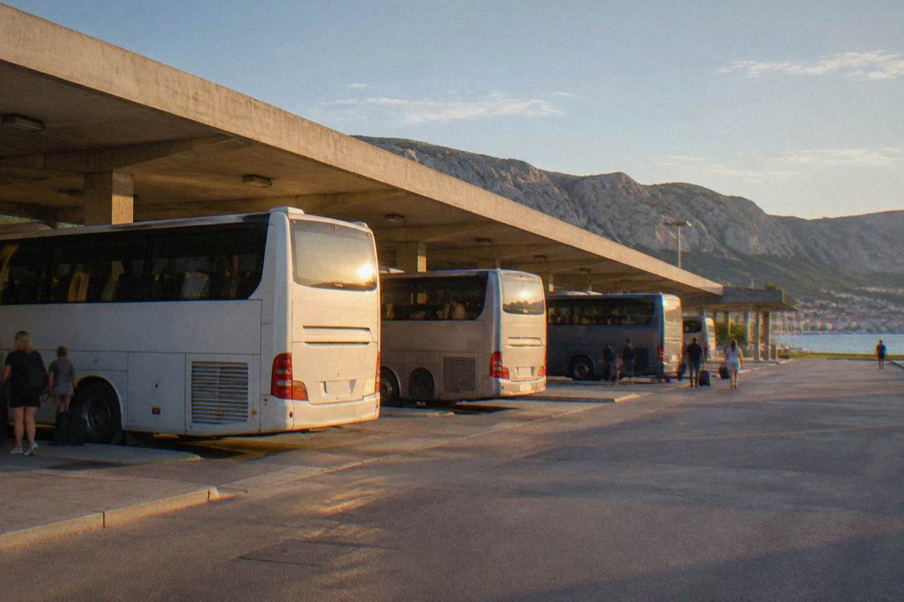 Coaches parked in a row beneath a plain concrete canopy with limestone hills rising behind