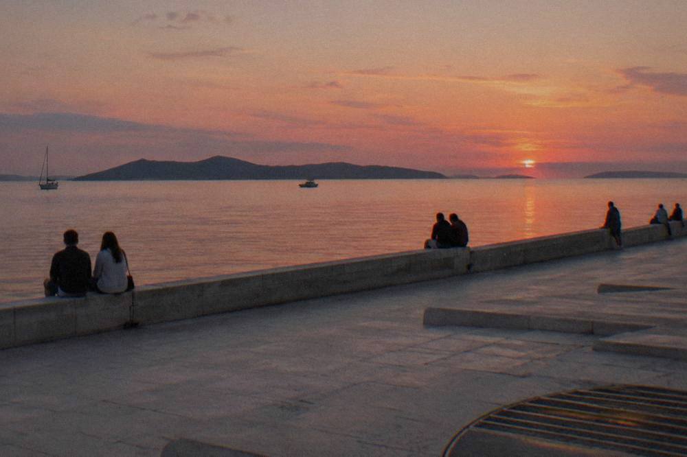 The stone seafront promenade at dusk with a few people at the wall facing a pink sky over the Adriatic