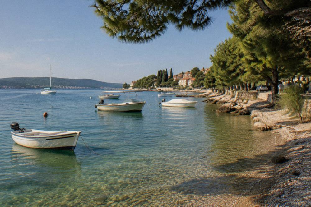 Boats anchored in clear shallow water by a pine-lined pebble beach on Ugljan island