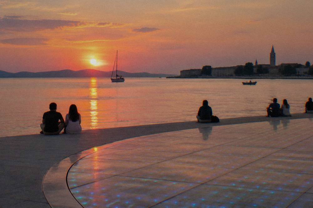 People seen from behind at the edge of a glowing circular installation by the sea at nightfall