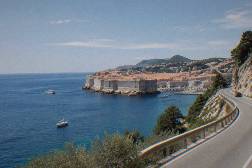 Coastal road winding past olive-green hills toward the sea in soft natural light
