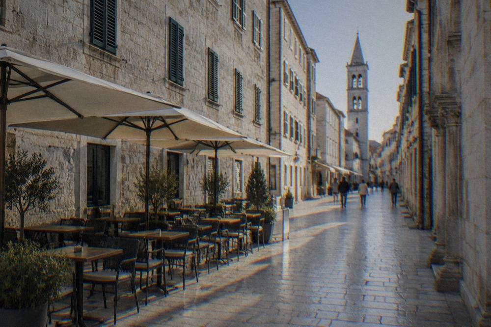 A pale limestone pedestrian street in Zadar Old Town with a quiet cafe terrace in morning light.