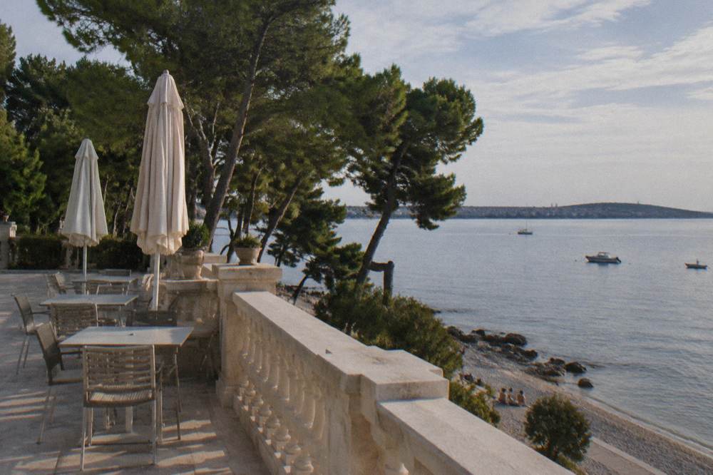 A quiet seaside hotel terrace with empty chairs overlooking a pine-fringed shore in Borik.