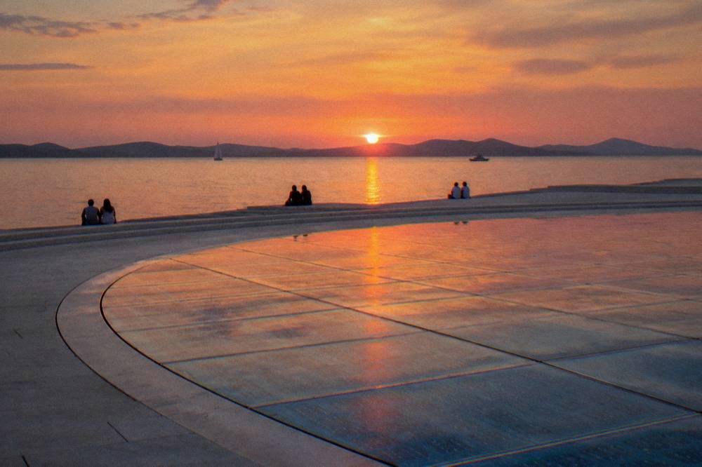 Distant people seated on pale waterfront steps at sunset beside the glass circles of the Greeting to the Sun.