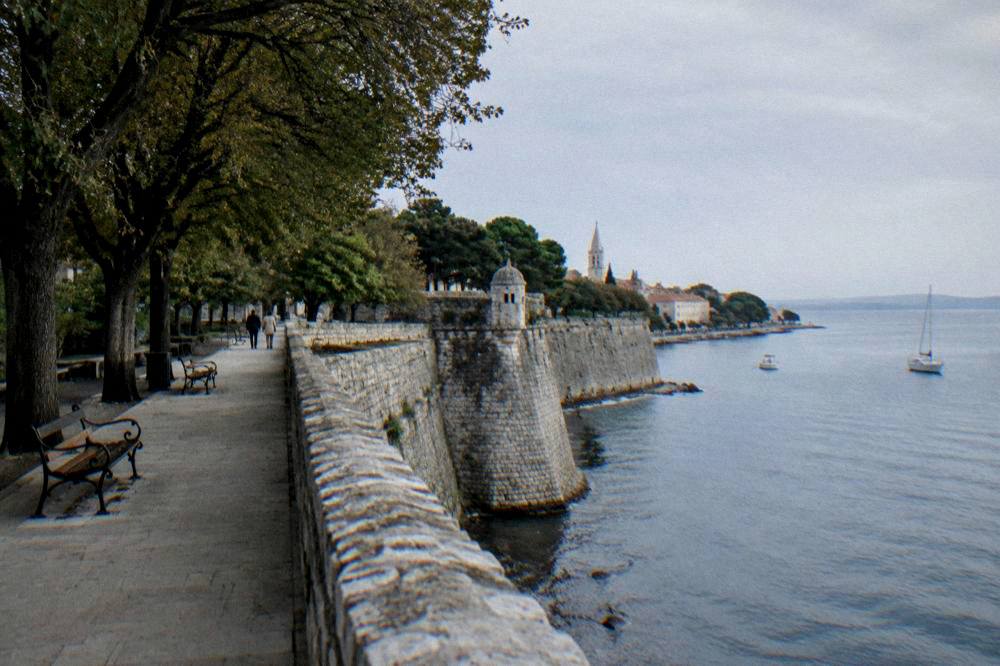 The stone ramparts of Zadar with a tree-lined promenade and empty benches above the sea.