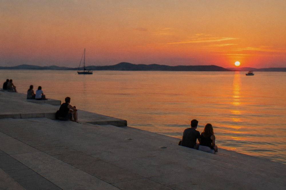 A few distant people on stone waterfront steps at sunset beside a calm sea.
