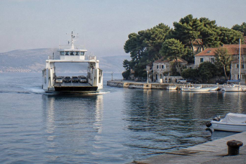 A white ferry approaching a small island harbour backed by pines and stone houses.