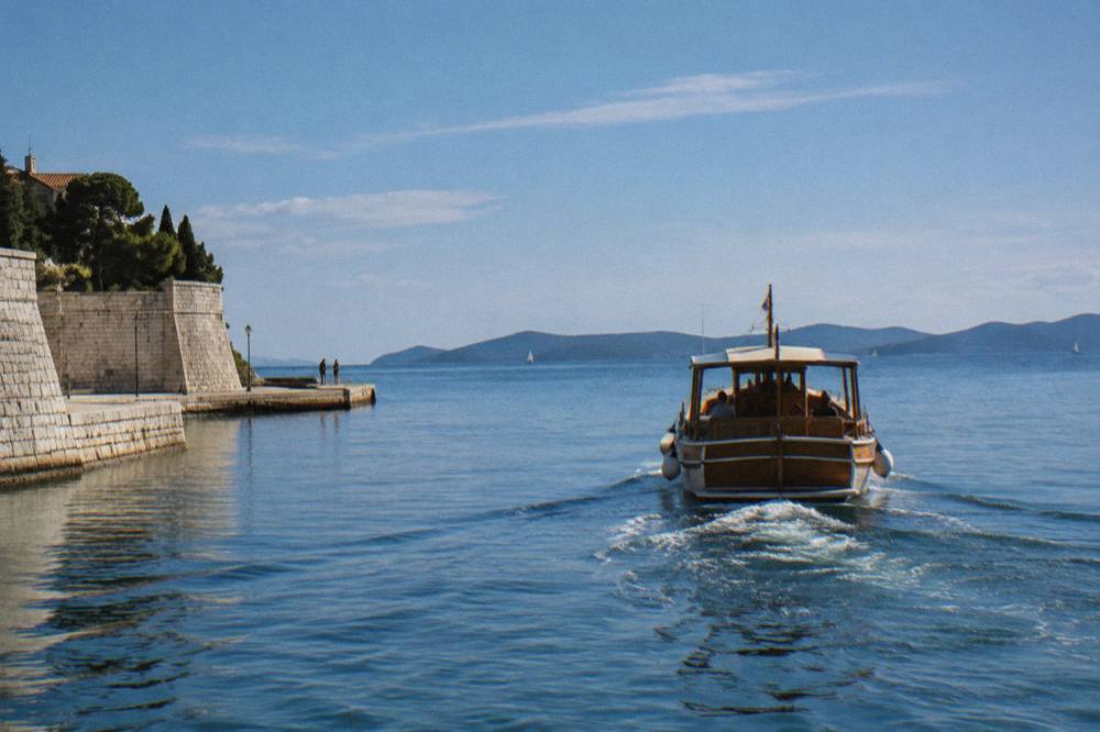 A wooden boat leaving the stone-walled Foša harbour toward islands on the horizon.
