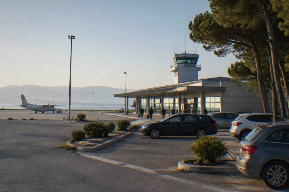 A small coastal airport terminal under a clear sky with parked cars in front.