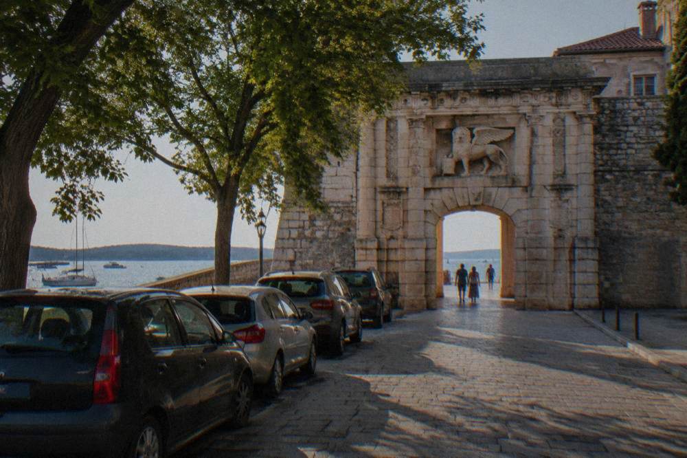 Cars parked beside the stone arch of the Land Gate in Zadar, shaded by trees.