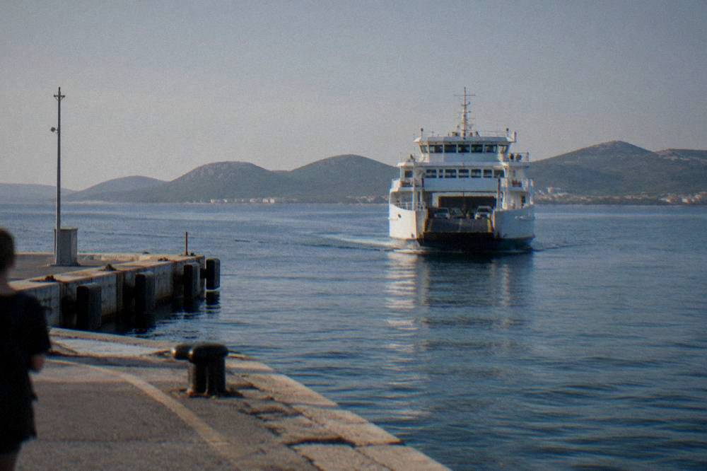 A car ferry approaching a dock with island hills behind it on calm blue water.