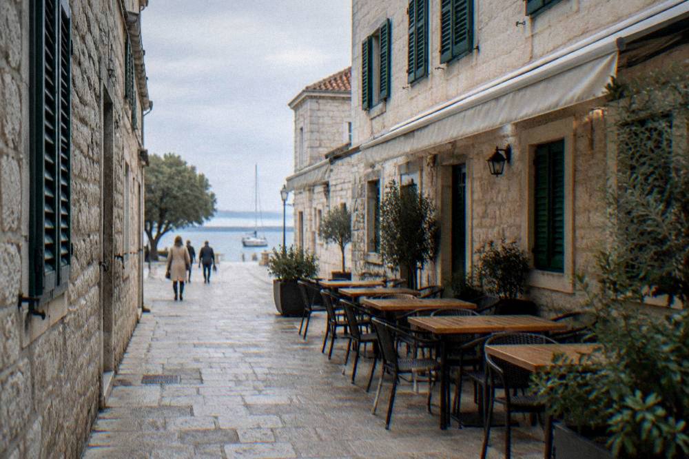 A narrow pale stone lane in Zadar Old Town with green shutters and empty cafe tables.