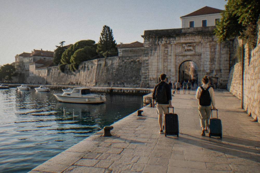 Two travellers with suitcases seen from behind walking along a sunlit waterfront toward the Sea Gate.