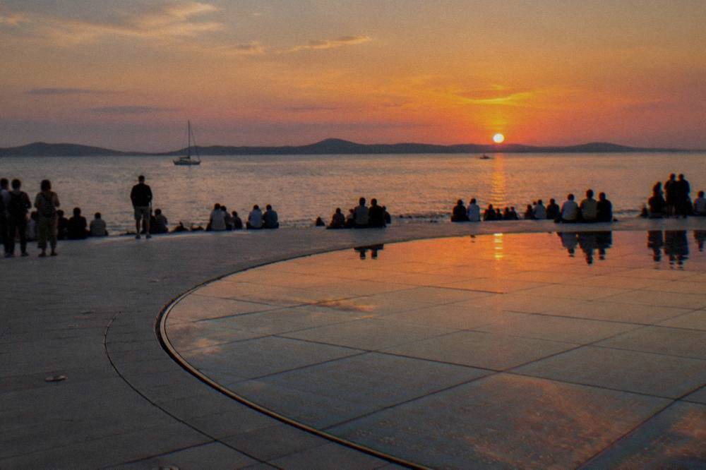 A distant crowd on waterfront steps watching an orange sunset beside a glass solar circle.