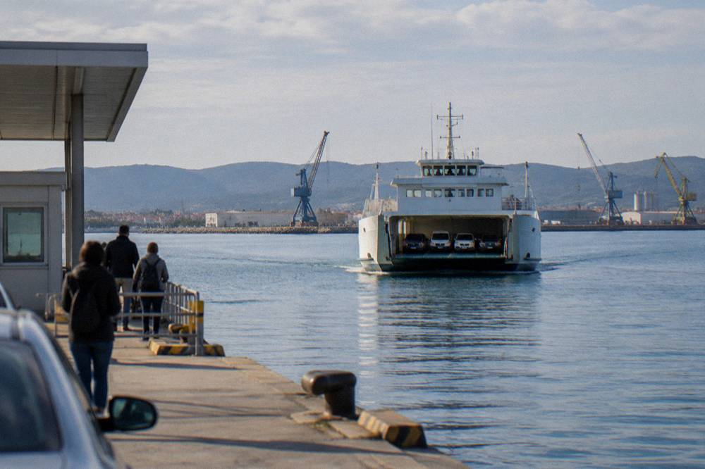 A car ferry approaching a port terminal with cranes and low hills behind.