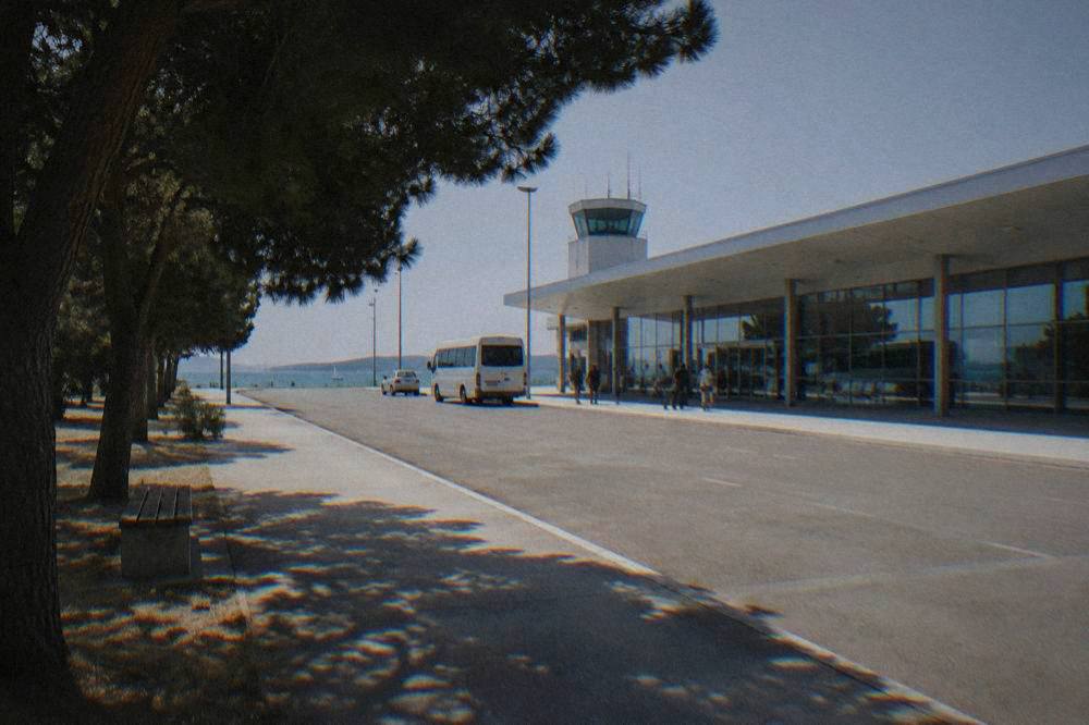 The arrivals forecourt of a small airport with an empty kerb in bright light.