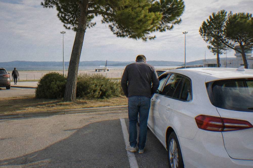 A driver seen from behind checking a white car in an airport car park.