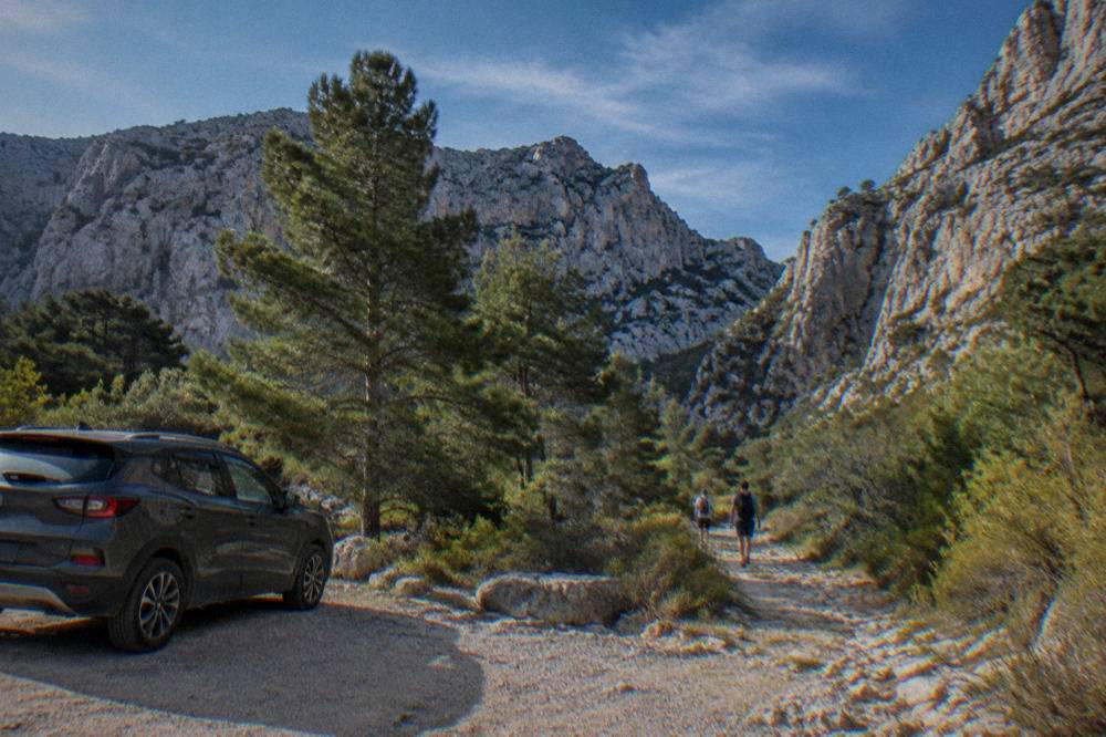 A car parked at a trailhead below steep grey limestone cliffs.