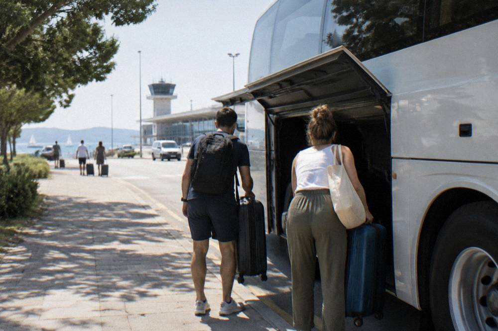 Two travellers seen from behind lifting suitcases into the luggage bay of a coach.