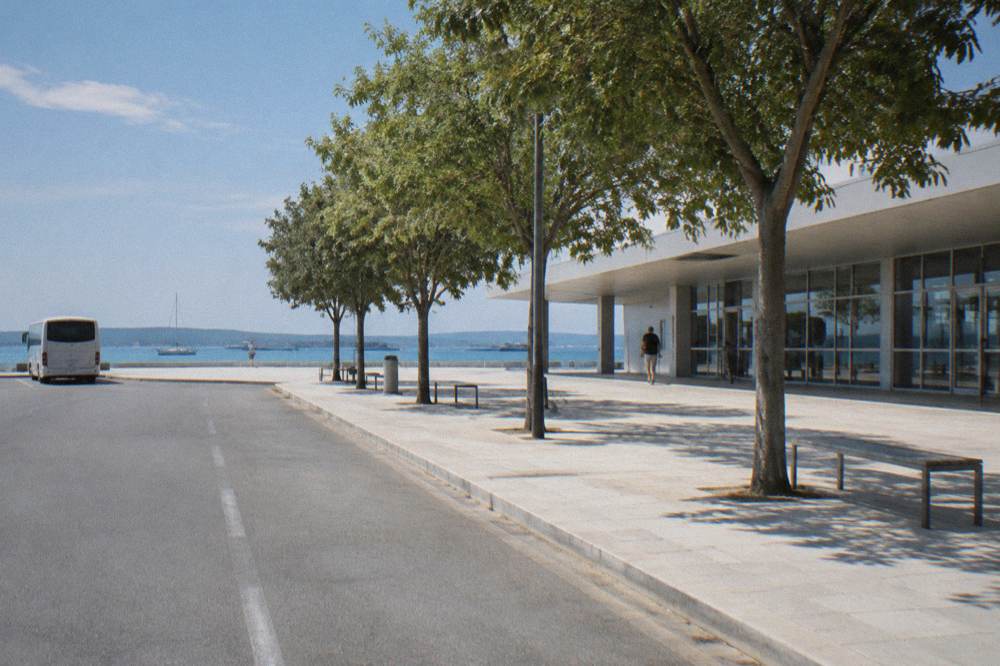 An empty kerb outside a modern building with trees, empty benches and pale paving.