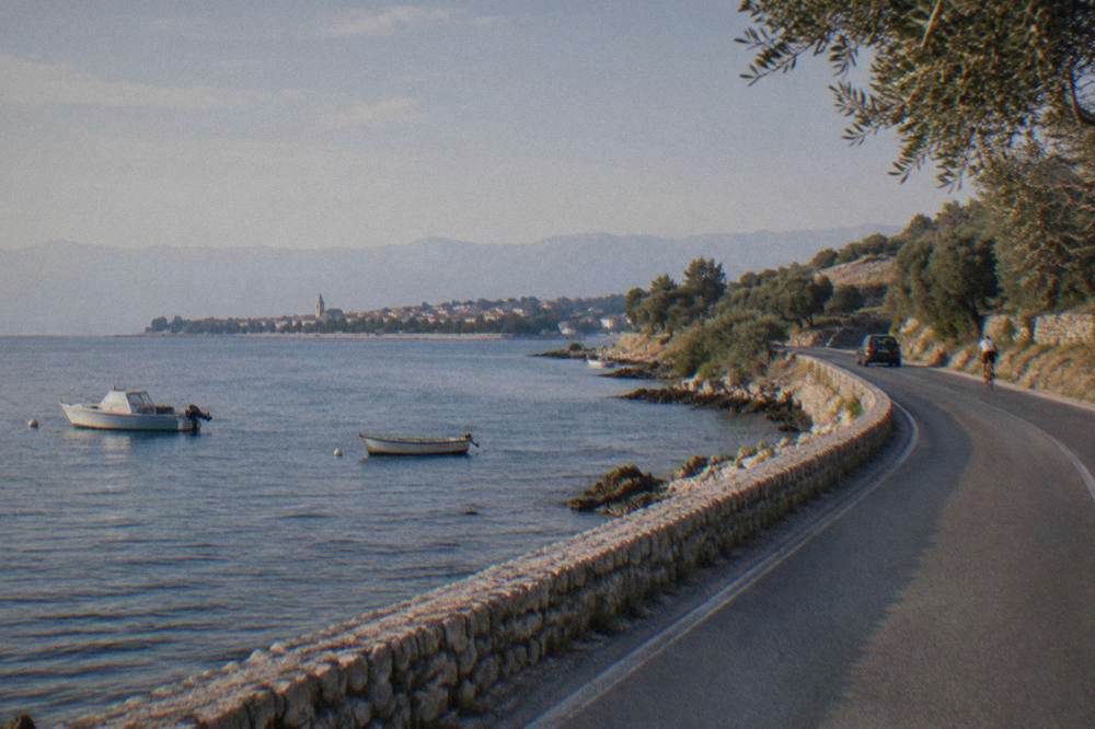 A coastal road curving along the Adriatic shoreline with dry hills and olive trees.