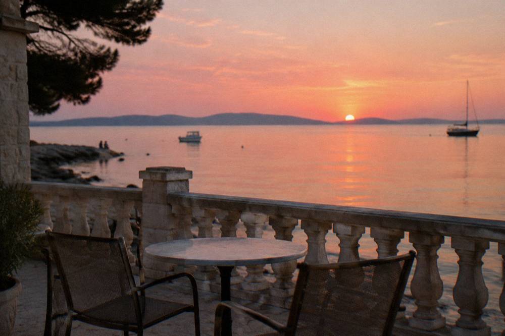 Sunset over the Adriatic from a terrace with a stone balustrade and empty chairs.