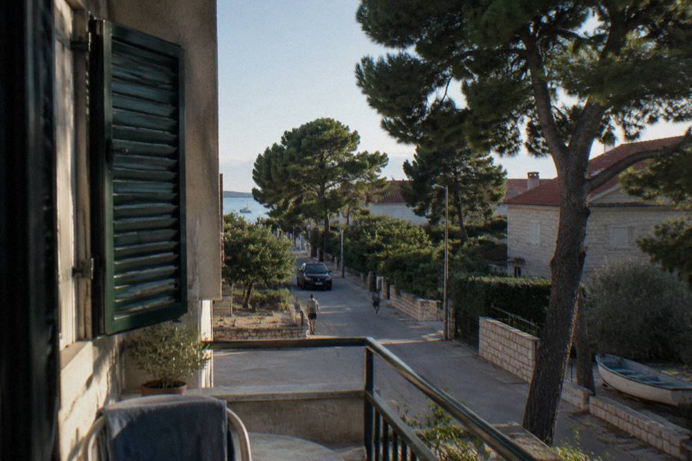 A balcony view over a quiet residential street with pines and low stone houses.