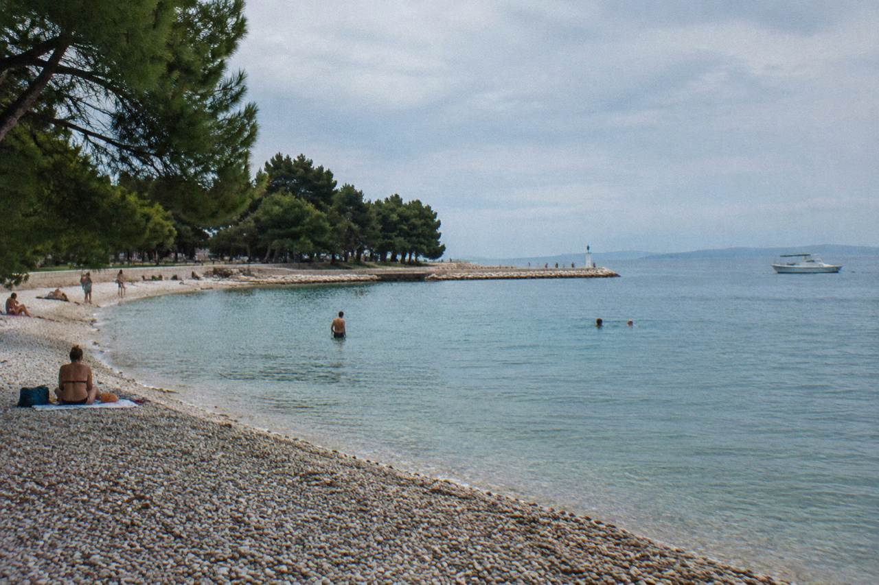 A pebble beach on Zadar's waterfront with calm turquoise water, pines behind.