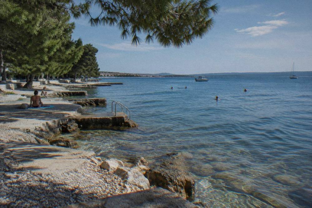 Concrete and pebble bathing platforms along a pine-shaded shore with clear water.