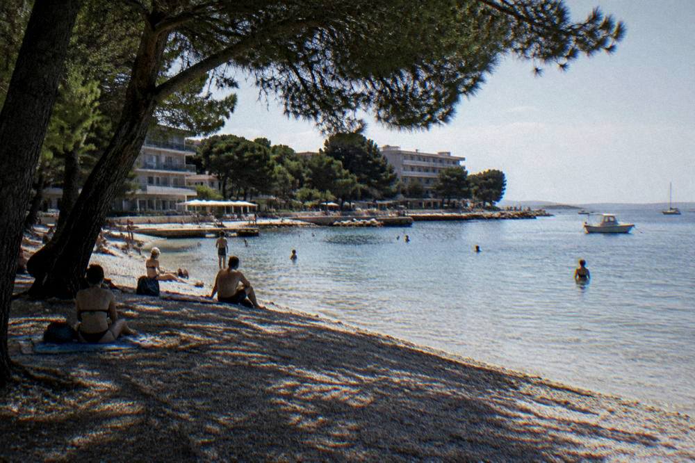 Bathers on a gravel beach in front of low seaside hotels under pines.